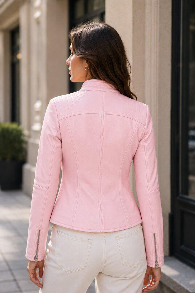 Puede incluir: Una chaqueta de color rosa claro con cuello alzado, paneles acolchados en los hombros y mangas largas con cremalleras met&aacute;licas plateadas en los pu&ntilde;os. La prenda presenta costuras verticales en la espalda y se combina con pantalones claros.