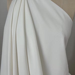 May include: A draped, off-white fabric is displayed on a mannequin. The fabric is folded and gathered, creating soft, flowing lines. The material appears to be smooth and opaque, with a subtle sheen. The overall impression is of elegance and simplicity.