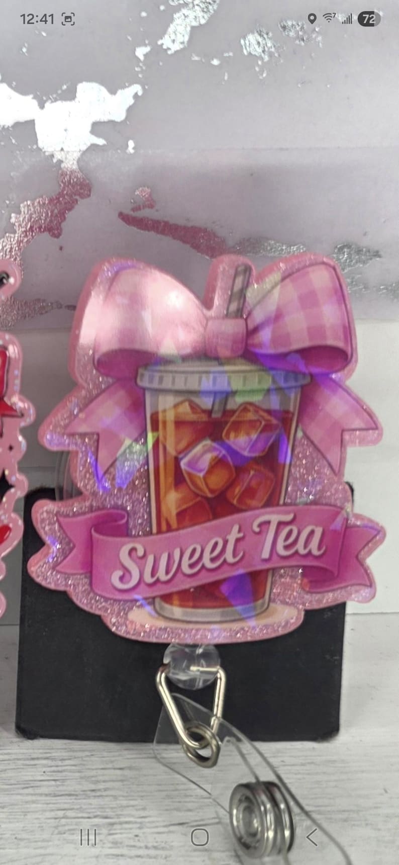 May include: A pink glitter badge reel featuring a sweet tea design. The design includes a cup of iced tea with ice cubes, a pink bow, and a banner that reads "Sweet Tea". The badge reel has a clear plastic retractable cord.