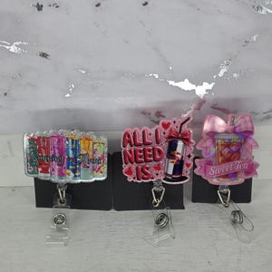 May include: Three badge reels with different designs. One says "Running on Mom" with colorful cans. Another says "All I Need Is" with a Red Bull can. The last one says "Sweet Tea" with a sweet tea graphic.