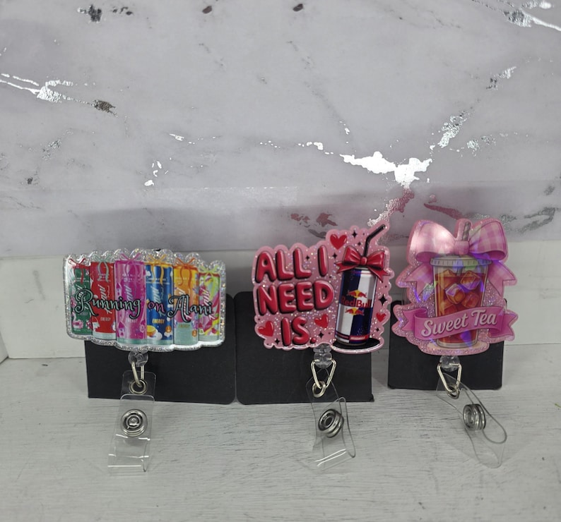 May include: Three badge reels with different designs. One says "Running on Mom" with colorful cans. Another says "All I Need Is" with a Red Bull can. The last one says "Sweet Tea" with a sweet tea graphic.