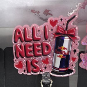 May include: A pink badge reel with the phrase "ALL I NEED IS" in red letters. The design includes a Red Bull can with a red bow, hearts, and glitter accents. The reel has a clear plastic strap and a metal clip.