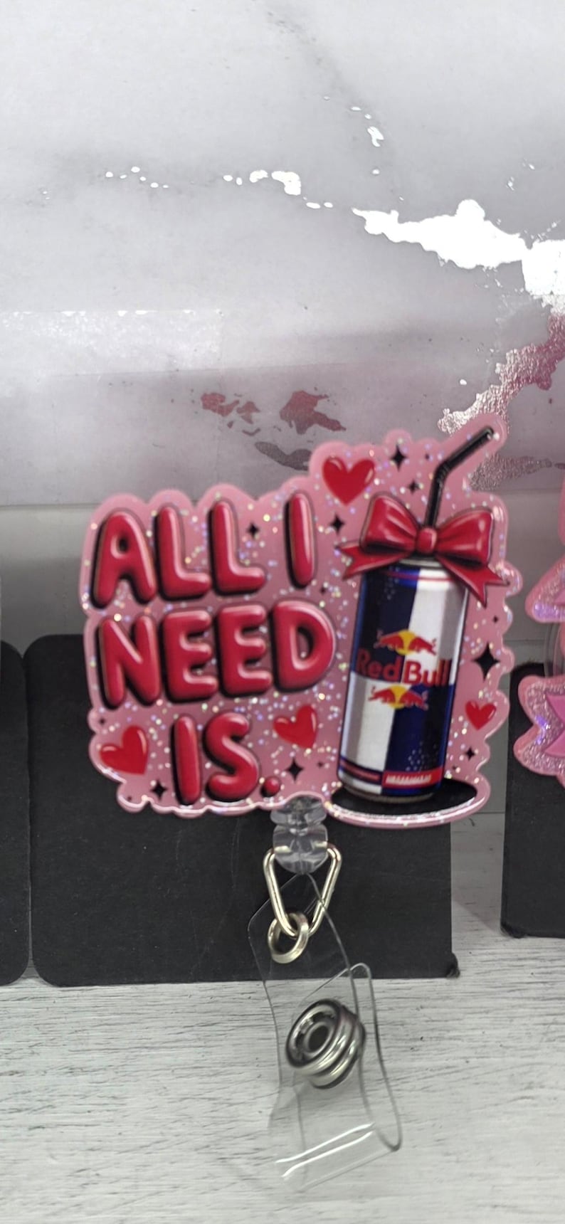 May include: A pink badge reel with the phrase "ALL I NEED IS" in red letters. The design includes a Red Bull can with a red bow, hearts, and glitter accents. The reel has a clear plastic strap and a metal clip.