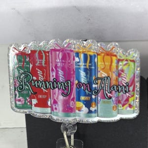 May include: A badge reel with a clear plastic strap and a metal clip. The badge reel has a rectangular design with the words "Running on Alani" and images of colorful energy drink cans.