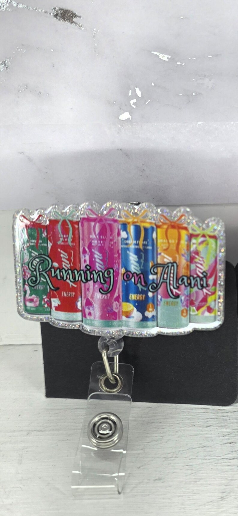 May include: A badge reel with a clear plastic strap and a metal clip. The badge reel has a rectangular design with the words "Running on Alani" and images of colorful energy drink cans.