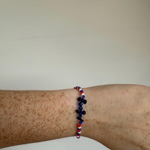 May include: A beaded bracelet with red, white, and blue beads, and a cluster of blue and black beads. The bracelet is worn on a freckled arm.