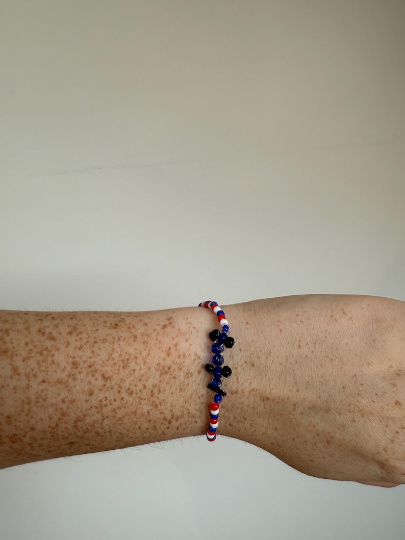 May include: A beaded bracelet with red, white, and blue beads, and a cluster of blue and black beads. The bracelet is worn on a freckled arm.
