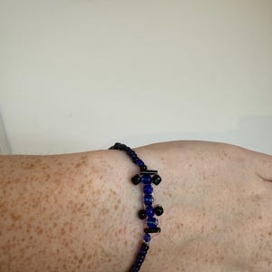 May include: A beaded bracelet featuring a central design of blue and black beads. The bracelet is composed of small, dark blue beads strung together. The central design has a series of larger blue beads and black accents.