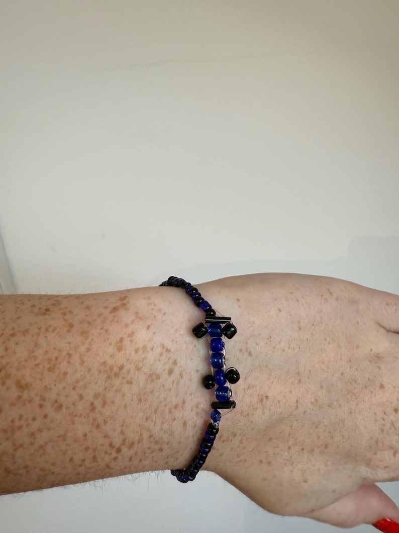 May include: A beaded bracelet featuring a central design of blue and black beads. The bracelet is composed of small, dark blue beads strung together. The central design has a series of larger blue beads and black accents.
