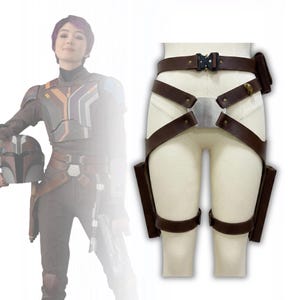 May include: Brown leather utility belt with two thigh holsters and a small pouch. The belt features a silver buckle and straps that cross the front. The belt is displayed on a mannequin.