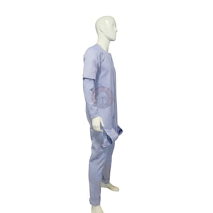 May include: A pale blue jumpsuit with long sleeves and a round neckline. The jumpsuit has a relaxed fit and features a pocket on the right side. The model is wearing white shoes. The jumpsuit is made of a soft, lightweight fabric.