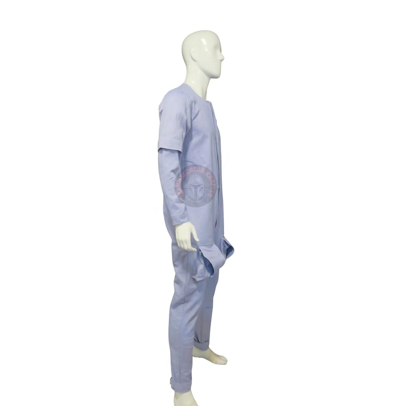 May include: A pale blue jumpsuit with long sleeves and a round neckline. The jumpsuit has a relaxed fit and features a pocket on the right side. The model is wearing white shoes. The jumpsuit is made of a soft, lightweight fabric.
