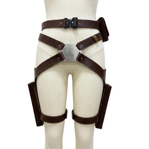 May include: Brown leather utility belt with multiple straps, pouches, and a metal buckle. The belt features a silver metal plate and is designed to be worn around the waist and thighs. The belt is displayed on a mannequin.