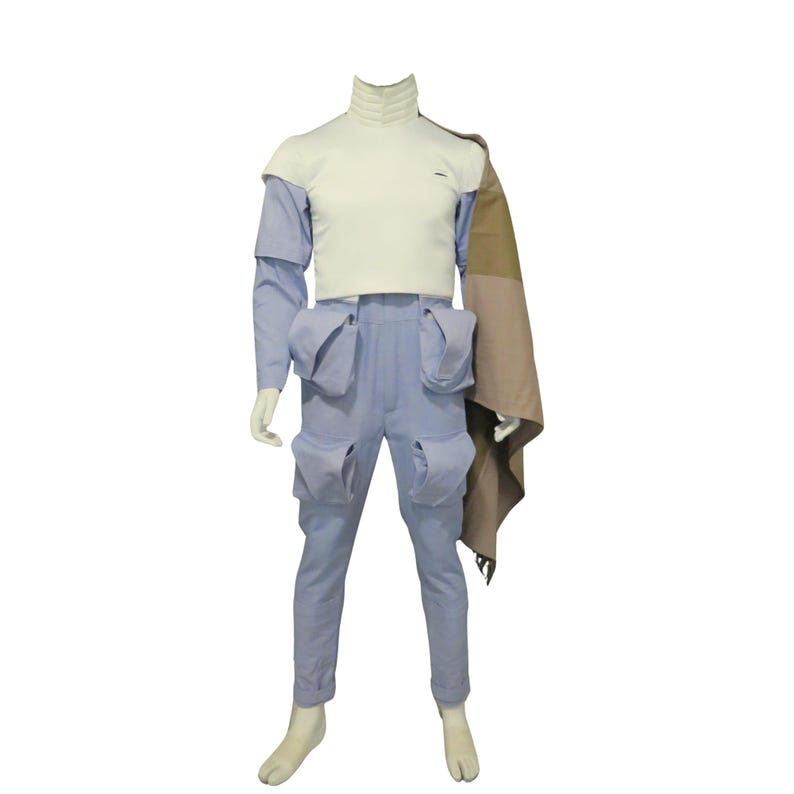 May include: A mannequin displaying a futuristic outfit. The outfit features light blue trousers with large pockets, a white high-necked top, and a cape draped over the shoulder. The ensemble is a mix of textures and colours.