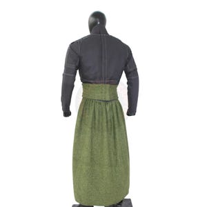 May include: A black jacket with long sleeves and a high collar, paired with a long, olive green skirt. The skirt has a high waist and a textured appearance. The outfit is displayed on a mannequin.