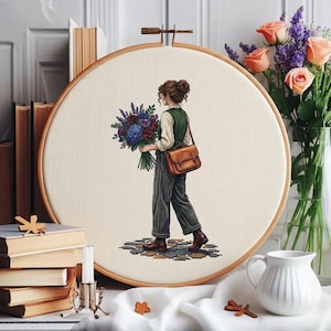 May include: A circular embroidery hoop containing a cross-stitch design of a person seen from behind, wearing a green vest, light shirt, and dark trousers, carrying a large bouquet of blue and purple flowers and a brown leather satchel.