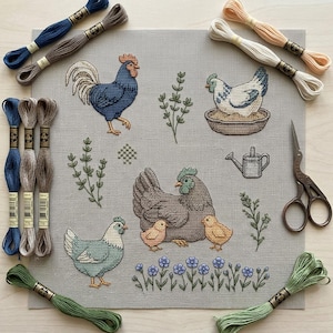 Puede incluir: Una pieza cuadrada de tela de lino gris con un patrón de punto de cruz de tres gallinas, dos pollitos, una regadera y varias plantas. El bordado está rodeado por varias madejas de hilo de bordar de colores y unas tijeras de metal.