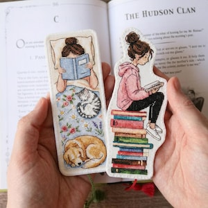 Puede incluir: Dos marcapáginas bordados con ilustraciones de una persona leyendo. Uno muestra a una persona leyendo en la cama con un gato y un perro. El otro muestra a una persona leyendo mientras está sentada en una pila de libros. Los marcapáginas están en manos de una persona.