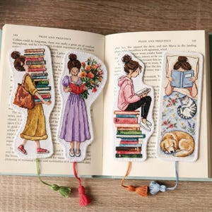 Op de afbeelding: Vier geborduurde bladwijzers met illustraties van lezende vrouwen. Eén vrouw draagt boeken, een andere houdt bloemen vast, een derde zit op boeken en de laatste leest in bed met een kat en een hond. Elke bladwijzer heeft een kwast.