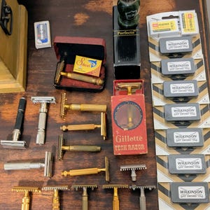 May include: A collection of vintage shaving tools, including safety razors, a shaving brush, and razor blades. The razors are in various styles and finishes, including gold and silver tones. The image also shows boxes of Wilkinson Sword razor blades.