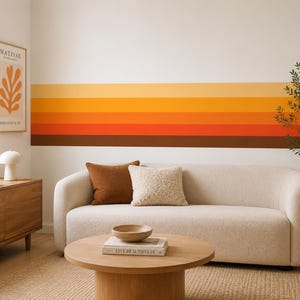 May include: A cream-coloured sofa with a brown square cushion and a textured cream cushion. Behind the sofa, a wall features horizontal stripes in shades of yellow, orange, red, and brown. A wooden coffee table sits in front with a book and bowl.
