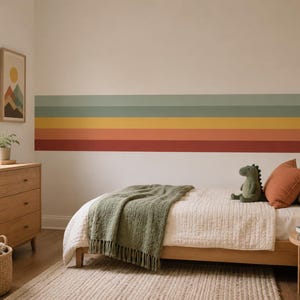 Op de afbeelding: Een slaapkamer met een houten bedframe, een wit dekbed, een textuurrijke saliegroene sprei en een pluche groene dinosaurus. De muur heeft horizontale strepen in saliegroen, geel en terracotta, passend bij de rest van het interieur.