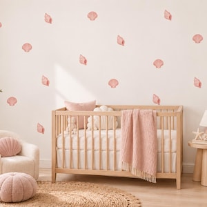 May include: A set of pink seashell wall decals arranged on a plain wall above a wooden crib. The crib features a white mattress, a pink knit throw blanket, and a pink pillow. A matching pink seashell pillow sits on a small armchair nearby.
