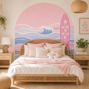 Könnte beinhalten: Ein Wandaufkleber mit einem rosa Bogen, stilisierten blauen und weißen Meereswellen und einem rosa Surfbrett mit weißen Blumenmustern. Das Design befindet sich hinter einem Bett mit Rattan-Kopfteil, rosa Kissen und einer weißen Decke.