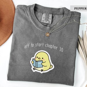 May include: A pepper-colored t-shirt featuring a graphic of a yellow chick reading a blue book. Above the chick, the text reads "off to start chapter 30" in white. The shirt has a crew neckline and a Comfort Colors brand label at the inner collar.