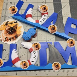 May include: Blue 3D-printed letters spelling ORCA, featuring white life preservers with red accents, grey shark fins, and circular stickers showing a cat with the text Huayra Crafts. The items are arranged on a cutting mat with inch markings.
