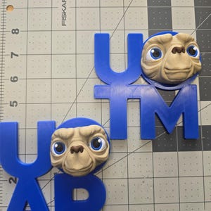 May include: Blue 3D-printed letters spelling out "UTM" and "UAP" with an E.T. character head attached to each. The E.T. heads are tan with large blue eyes and a blue head. The letters are on a gray cutting mat.
