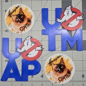 May include: A collection of stickers and blue 3D-printed letters. The stickers feature a cat illustration and the text "Huayra Crafts". Two stickers feature a Ghostbusters logo. The blue letters spell out "YAP" and "YTM".