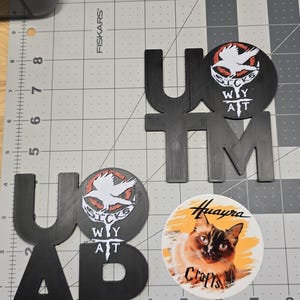 May include: Black wooden letters spelling out "UAP" and "UTM" are arranged on a gray cutting mat. Two round stickers feature a white bird design with text. A round sticker with a cat image and the text "Huayra Crafts" is also present.