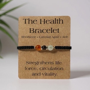 May include: A black bracelet with three gemstone beads: orange, black, and green. The bracelet is displayed on a brown card that reads "The Health Bracelet" and lists the gemstones. The card also states the bracelet strengthens life force, circulation, and vitality.