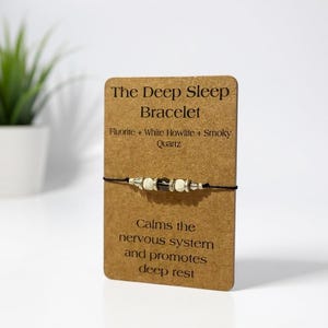 May include: A bracelet with Fluorite, White Howlite, and Smoky Quartz beads is displayed on a brown card. The card reads "The Deep Sleep Bracelet" and includes text about calming the nervous system and promoting deep rest. A small potted plant is in the background.