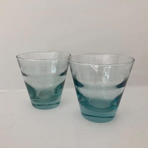 May include: Two matching drinking glasses made of translucent light blue glass. Each glass features a wide, open top that tapers down to a narrower, circular base. The glass material is smooth and clear, showing a subtle tint throughout the body.