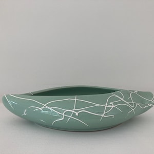 May include: A boat-shaped ceramic bowl in a soft sage green colour, featuring a glossy finish and a decorative white drip pattern that crisscrosses across the exterior surface. The vessel has a smooth, elongated, and curved silhouette.