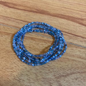 May include: A multi-strand bracelet composed of small, circular glass beads in various shades of blue, including light sky blue, medium cobalt, and deep navy, with some translucent and opaque finishes arranged in a continuous circular loop.