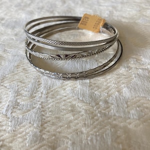 Peut inclure: Un ensemble de cinq bracelets joncs en métal argenté empilés. Les bracelets présentent des textures variées, notamment des motifs lisses, quadrillés et en corde torsadée. Une petite étiquette en papier beige est fixée au sommet.
