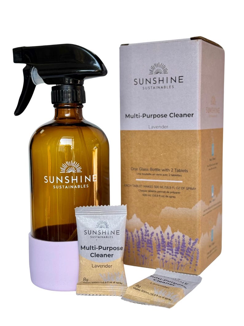 May include: Amber glass spray bottle with a black sprayer, a light purple base, and a cardboard box. The box and bottle feature the text "SUNSHINE SUSTAINABLES Multi-Purpose Cleaner Lavender". Two small packets of cleaner are also included.