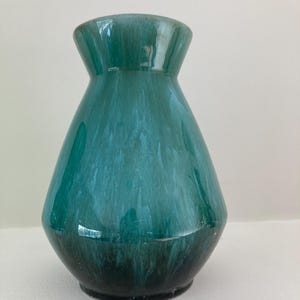 Peut inclure: Un vase en céramique brillant avec un col évasé et un corps fuselé, présentant un émail dégradé passant d'un bleu sarcelle foncé à la base à une teinte turquoise translucide plus claire vers le haut avec des stries blanches verticales.