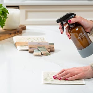May include: A brown glass spray bottle with a black nozzle and a gray silicone sleeve is being used to spray a white and green patterned cleaning cloth. The bottle has the text "SUNSHINE" on it. Other cleaning supplies are in the background.