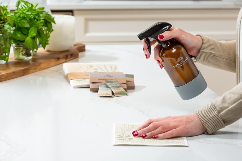 May include: A brown glass spray bottle with a black nozzle and a gray silicone sleeve is being used to spray a white and green patterned cleaning cloth. The bottle has the text "SUNSHINE" on it. Other cleaning supplies are in the background.