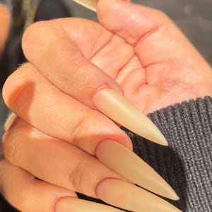 Könnte beinhalten: Eine Hand mit langen, mandelförmigen, beigefarbenen Acrylnägeln. Die Nägel sind einfarbig und haben ein mattes Finish. Die Hand ruht auf einem dunkelgrauen Pullover.