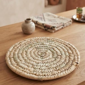 Puede incluir: Un mantel individual redondo y plano tejido con hojas de palma secas naturales en un patrón en espiral. El mantel presenta una paleta de colores beige claro y tostado con variaciones sutiles en la textura de las fibras tejidas.