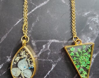May include: Two gold-colored necklaces with pendants containing dried flowers. One pendant is teardrop-shaped with a light blue flower, the other is a triangle with green flowers. Both have gold chains.