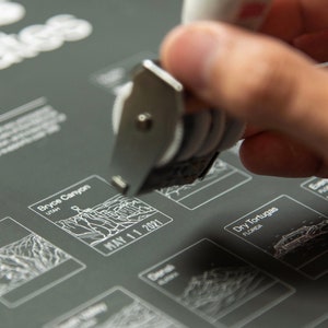 May include: A close-up of a hand using a date stamper on a gray poster with a list of national parks. The poster features the names of the parks, their locations, and a space to mark the date of a visit. The date stamper is being used to mark the date "MAY 11 2021" on the poster.