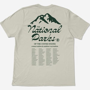 May include: A light beige t-shirt with a green mountain graphic and the words "The National Parks" in a cursive font. Below the text is a list of U.S. National Parks. The shirt promotes conservation and outdoor recreation.