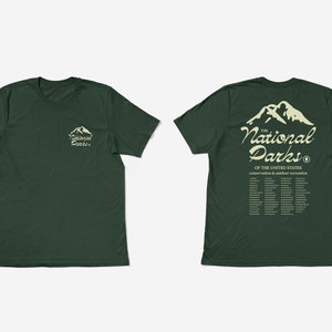 May include: Two dark green t-shirts. One shows a mountain graphic and the text "The National Parks" on the front and back. The back of the shirt also lists the names of the National Parks. The other shirt has the same mountain graphic and text on the front.