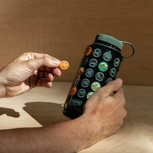May include: A black water bottle with a green lid and a handle. The bottle is covered in stickers of national parks. A hand is holding a sticker with a park logo.